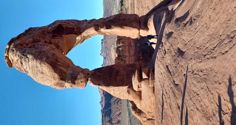 El icónico Arco Delicado elevándose sobre dos excursionistas en arenisca roja bajo la luz del atardecer.