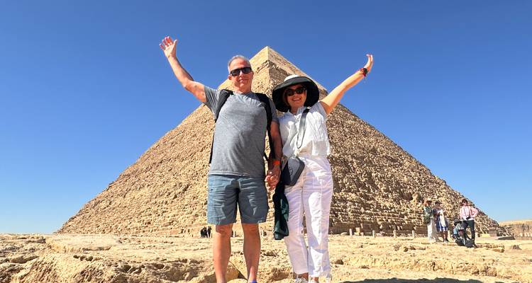 Pareja feliz levantando los brazos frente a la Pirámide de Kefrén bajo un cielo azul profundo.