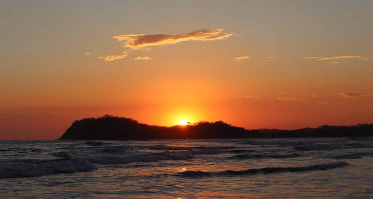 Sol naranja ardiente poniéndose detrás de un promontorio, iluminando suaves olas en una playa tranquila.