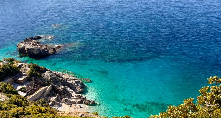 Cala turquesa del Adriático con agua cristalina, afloramientos rocosos y pequeña playa escondida vista desde arriba.