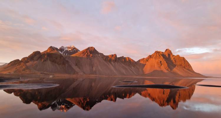 Luz dramática de amanecer naranja reflejando montañas escarpadas en las aguas quietas de la laguna en Vestrahorn
