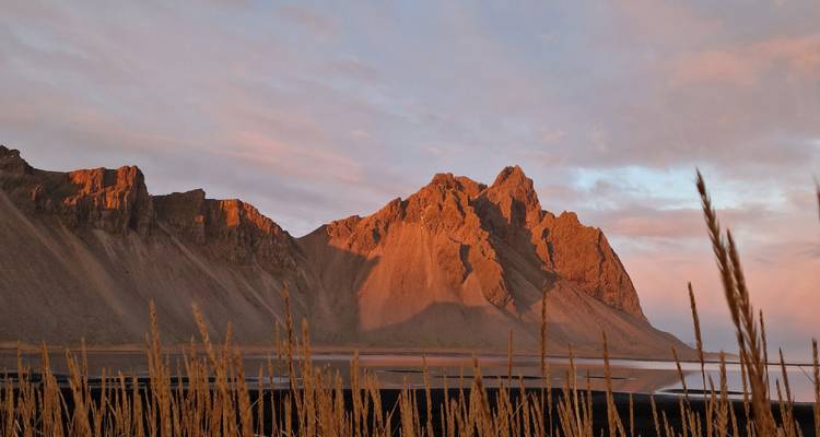 Dramáticos picos montañosos brillan en color naranja bajo el sol bajo, enmarcados por pastos altos y una ensenada tranquila.