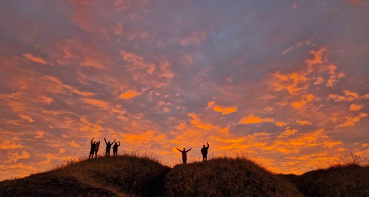 Siluetas de amigos celebran sobre dunas cubiertas de hierba contra un cielo dramático de atardecer ardiente.
