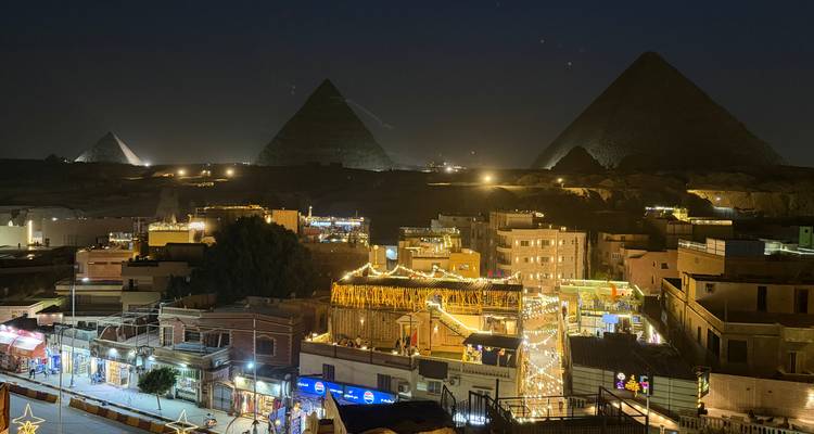 Nachtpanorama der beleuchteten Pyramiden von Gizeh, die sich über ein lebendiges, von Basaren beleuchtetes Viertel erheben.
