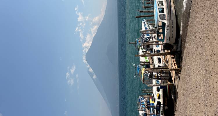 Clear view of small boats moored at a lakeside pier with a prominent volcano rising across turquoise waters under a blue sky.