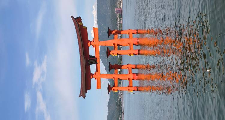 Le célèbre portail torii d'un rouge vermillon éclatant du sanctuaire d'Itsukushima se dressant dans les eaux calmes et bleues de la mer, avec les montagnes lointaines et la ville sur le rivage.