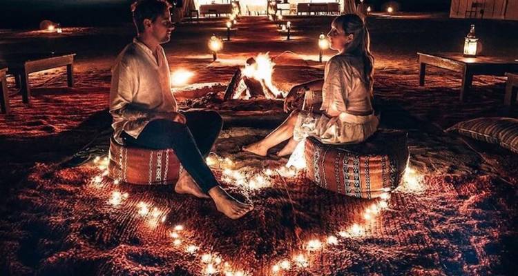 Couple seated on cushions inside a heart-shaped ring of fairy lights beside a campfire at a desert glamping site at night.