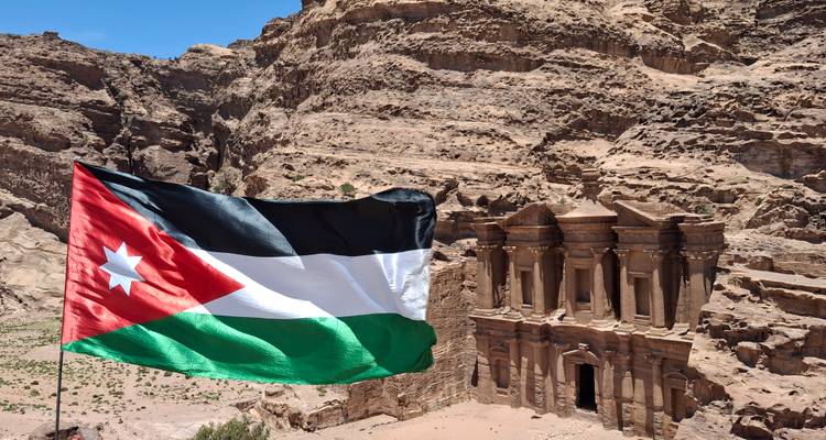 La bandera jordana ondea en primer plano con vistas al majestuoso Monasterio de Petra y los acantilados circundantes.
