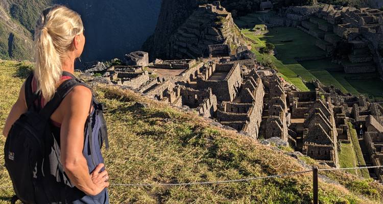 Una excursionista contempla las terrazas de piedra y las ruinas de Machu Picchu desde un mirador cubierto de hierba.