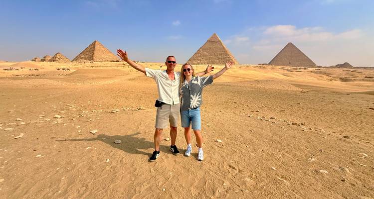 Pareja feliz posando con los brazos levantados frente a las Grandes Pirámides en el desierto arenoso
