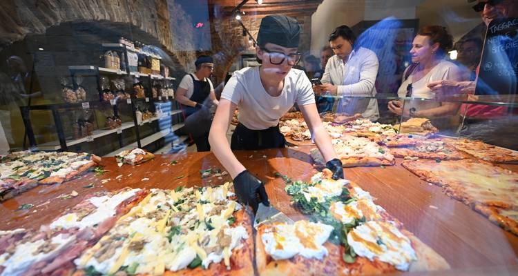 Le chef tranche des pizzas rectangulaires fraîchement préparées derrière un comptoir vitré tandis que les clients observent avec intérêt.