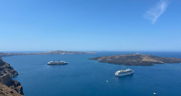 Vue aérienne large sur la caldeira de Santorin avec des navires de croisière ancrés dans les eaux bleu profond sous un ciel dégagé.