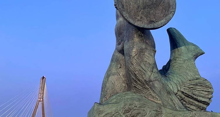 La icónica estatua de la sirena de Varsovia en bronce con el puente atirantado de fondo contra un cielo despejado.