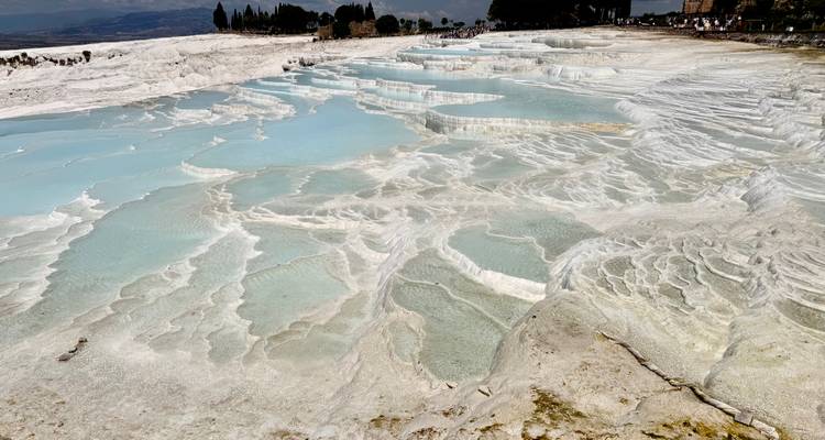 Melkwit-blauwe mineraalpools en witte travertijnen terrassen vallen in trappen naar beneden langs een helling onder de felle zon.