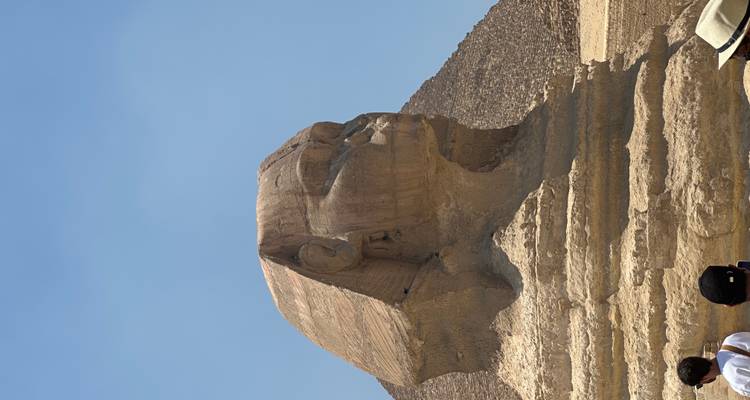 Grote Sphinx van Giza in profielzicht met piramidebasis erachter onder heldere hemel, toeristen eronder.