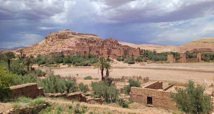 Panoramischer Blick auf das befestigte Lehmorf Ait Benhaddou, umgeben von Palmen und Wüstenhügeln unter einem dramatischen Himmel.