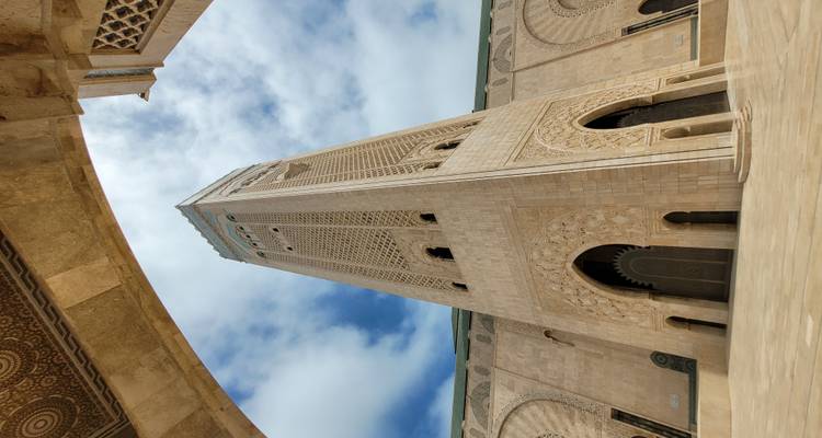 Aufwärtsansicht des kunstvollen Minaretts und der Arkaden der Hassan-II.-Moschee vor einem teilweise bewölkten blauen Himmel.