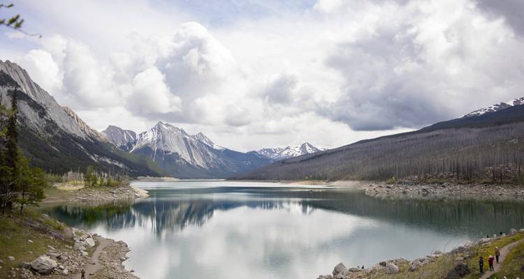 Lago sereno en la montaña que refleja picos nevados y nubes dramáticas con pequeños senderistas en la orilla.