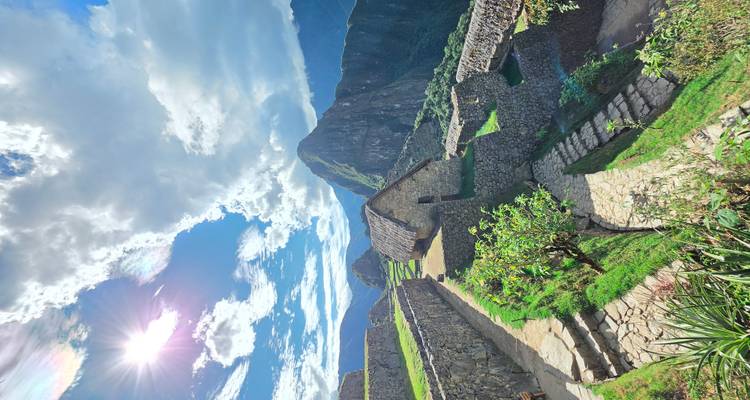 Sun-lit view of Machu Picchu terraces, stone houses and surrounding peaks beneath a dramatic sky.