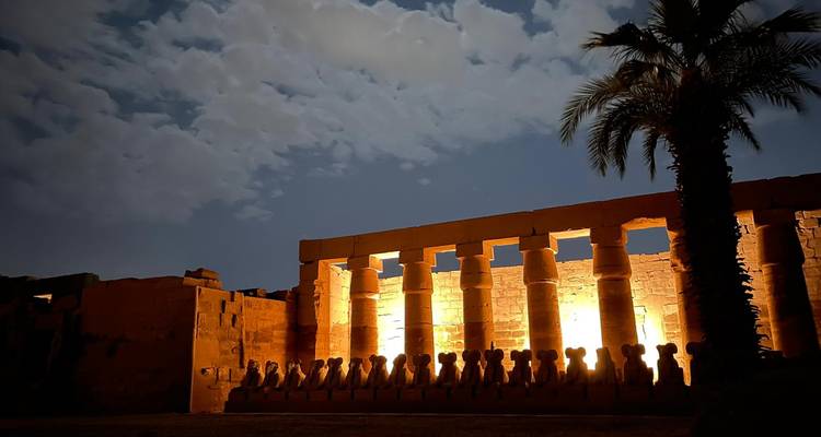 Night view of illuminated temple colonnade with dramatic lighting against a moonlit cloudy sky and palm silhouette.