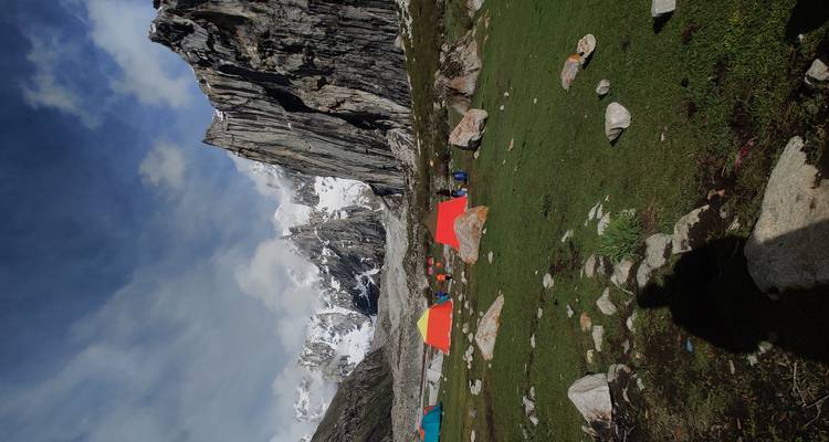 Des tentes d'expédition colorées parsemant un campement alpin herbeux sous des falaises imposantes striées de neige, sous un ciel spectaculaire.