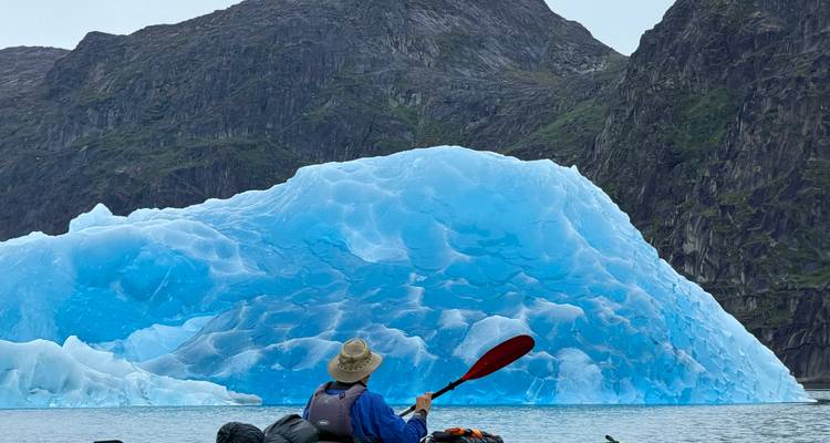 Un kayakista rema hacia un iceberg azul brillante recortado contra las empinadas montañas de Groenlandia.