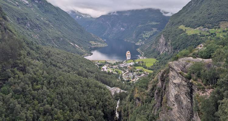 Vue panoramique sur le Geirangerfjord avec un navire de croisière, un village et des falaises verdoyantes luxuriantes sous de bas nuages.