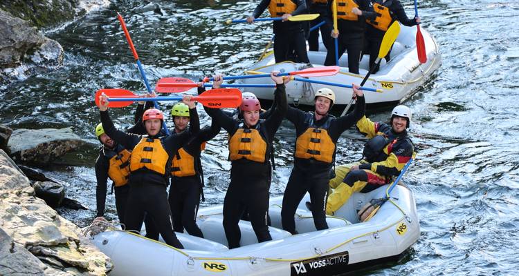 Groupe de canotiers en combinaisons de plongée qui crient de joie et lèvent les pagaies en naviguant sur une rivière aux courants rapides.