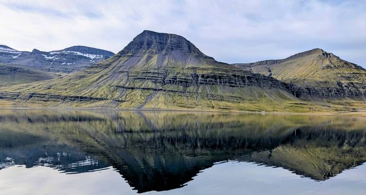 Zerklüfteter grüner Berg, der sich perfekt in einem ruhigen Fjord unter einem blassen Himmel in Island spiegelt
