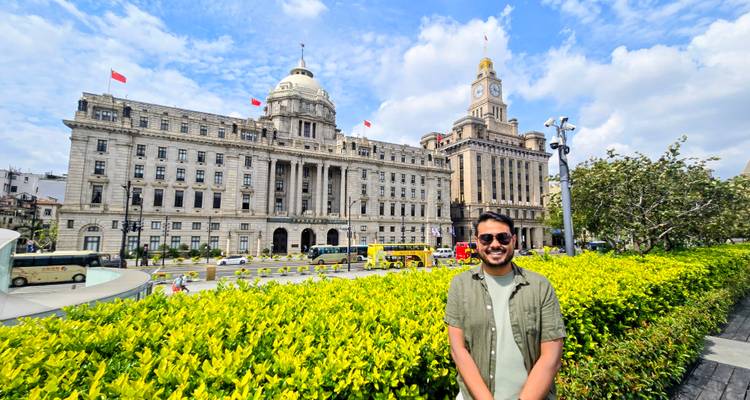 Touriste souriant se tenant devant les grands bâtiments coloniaux du Bund sous un ciel bleu et une verdure éclatante.