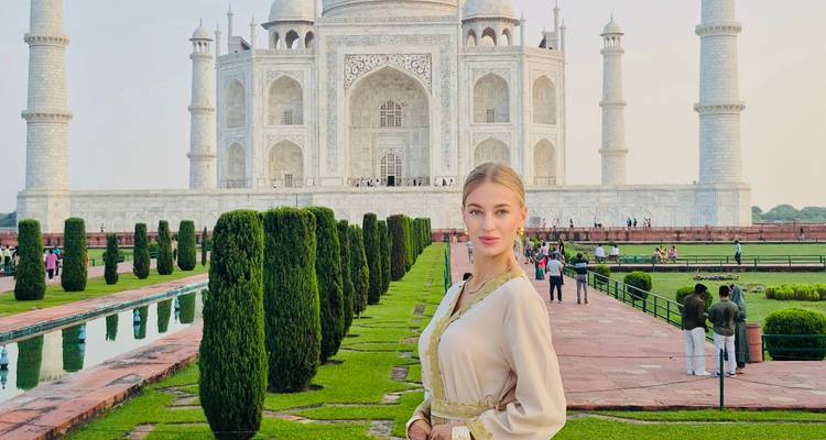 Mujer elegante posando junto al estanque de reflejos con el Taj Mahal elevándose detrás de ella al atardecer.