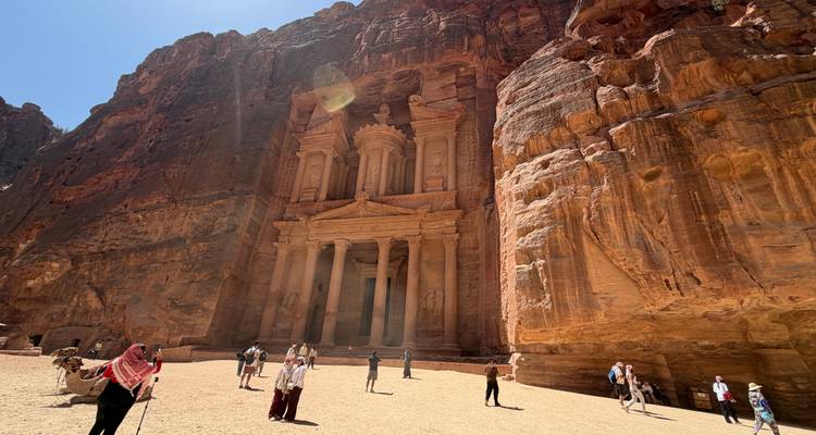 Sunlight streams onto the Treasury’s ornate façade at Petra as visitors explore the sandy plaza.