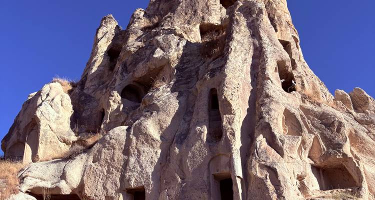 Une imposante formation rocheuse de couleur miel sculptée de habitations troglodytiques anciennes sur un ciel bleu clair.