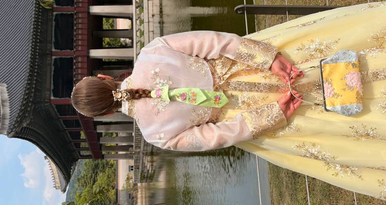 Une femme vêtue d'un élégant hanbok jaune se tient aux côtés d'un étang paisible du palais, un pavillon orné et un toit tuiled se dressant devant des collines verdoyantes.