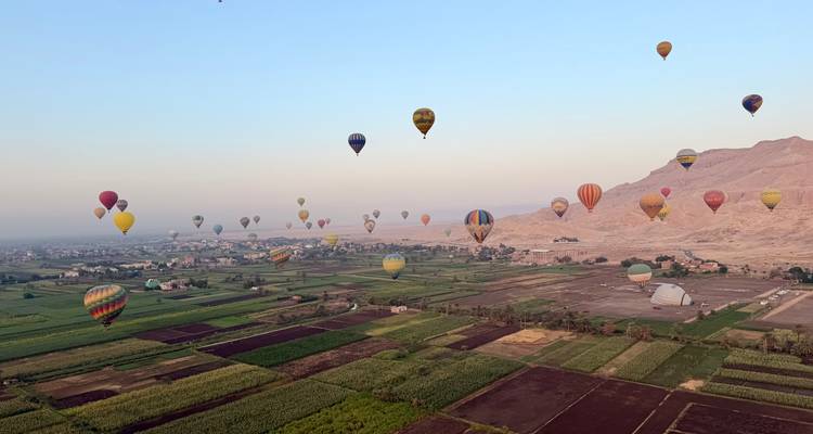 Des dizaines de montgolfières colorées flottant au-dessus de champs verts en damier et de collines désertiques à l'aube