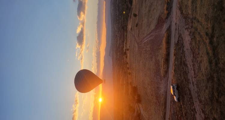Silhouette d'une montgolfière flottant au-dessus de la Cappadoce au lever du soleil avec un soleil doré à l'horizon et un véhicule isolé sur une piste de terre en dessous.