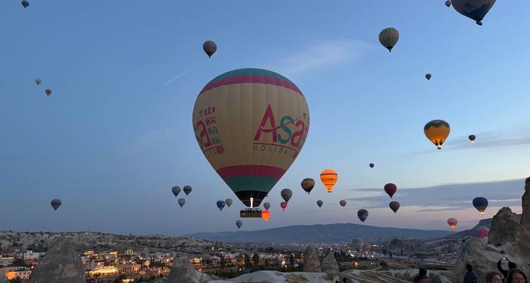 Docenas de coloridos globos aerostáticos se elevan sobre los valles de Capadocia al amanecer, con un globo prominente etiquetado como 'Asa'.
