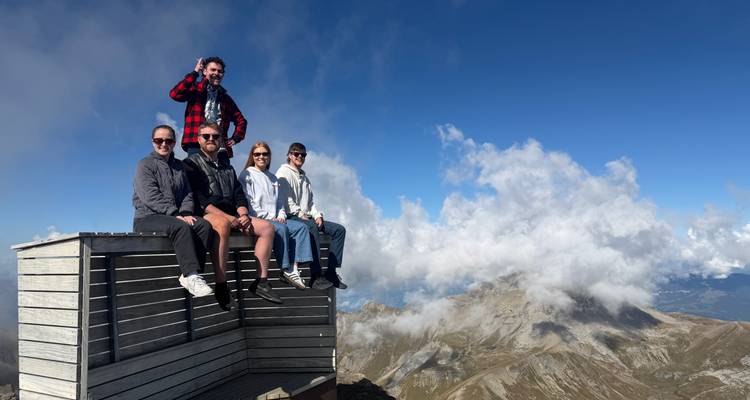 Reisende, die auf einer hölzernen Aussichtsplattform hoch in den Alpen sitzen, mit dramatischen Wolken und Bergspitzen im Hintergrund.