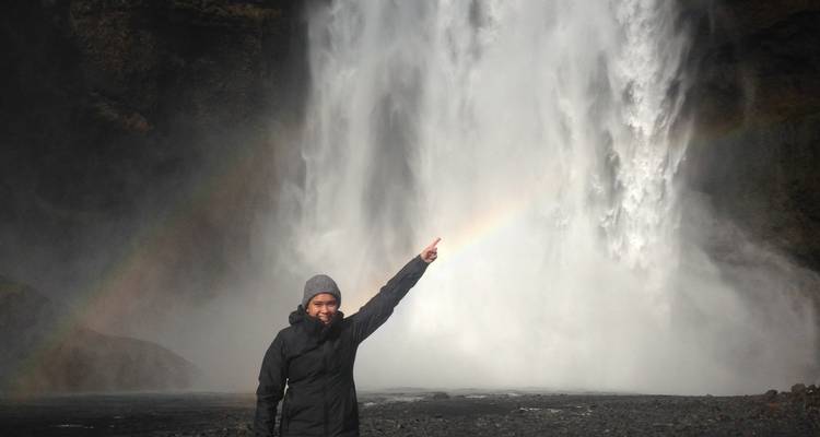 Person, die auf einen Wasserfall mit einem Regenbogen im Nebel zeigt.