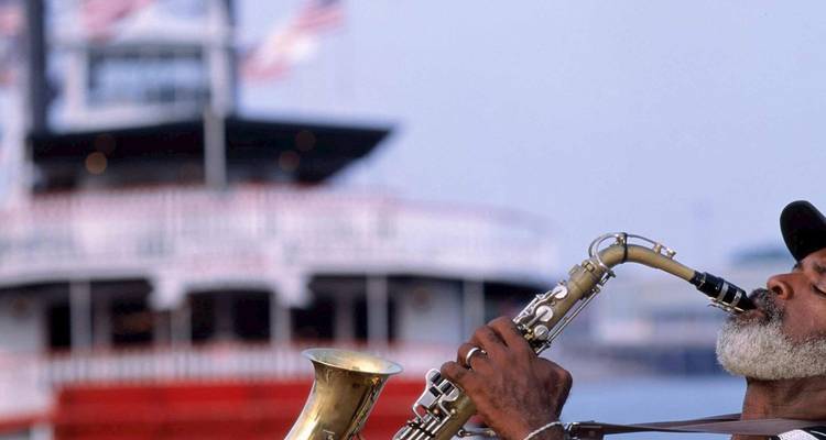Un músico callejero toca apasionadamente un saxofón en el río Mississippi con un barco de vapor de ruedas de paletas desenfocado al fondo