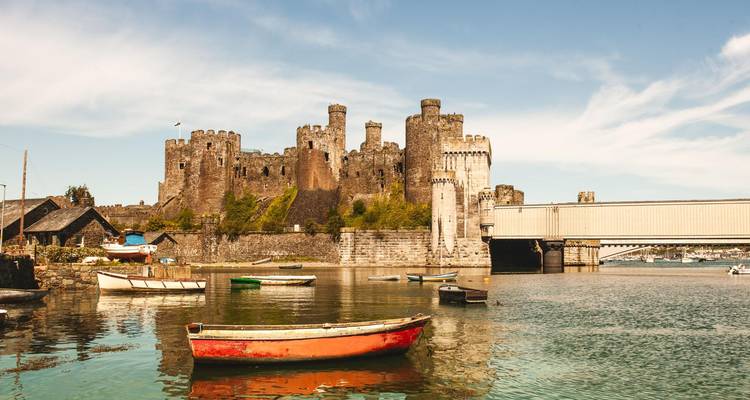 Le château médiéval en pierre de Conwy et le pont suspendu se reflètent dans la rivière calme avec de petits bateaux colorés.