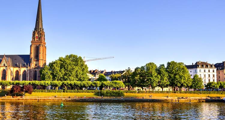 Panorama riverain de Francfort avec un haut clocher d'église gothique et une promenade verdoyante se reflétant dans le Main sous la lumière dorée du soir.