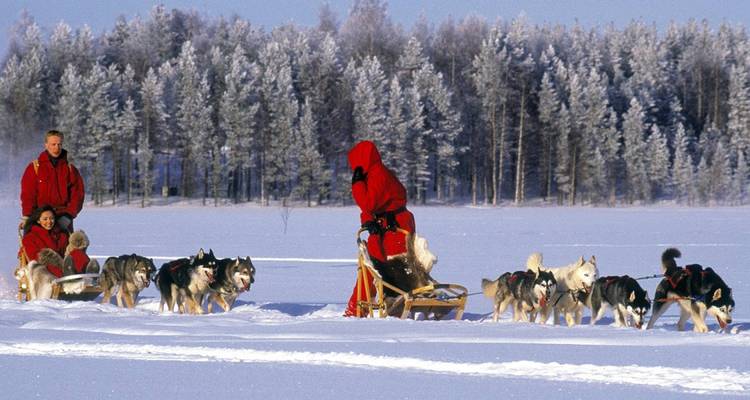 Une meute de chiens de traîneau huskies tire deux passagers à travers un vaste paysage enneigé tandis qu'un musher les guide de l'arrière.