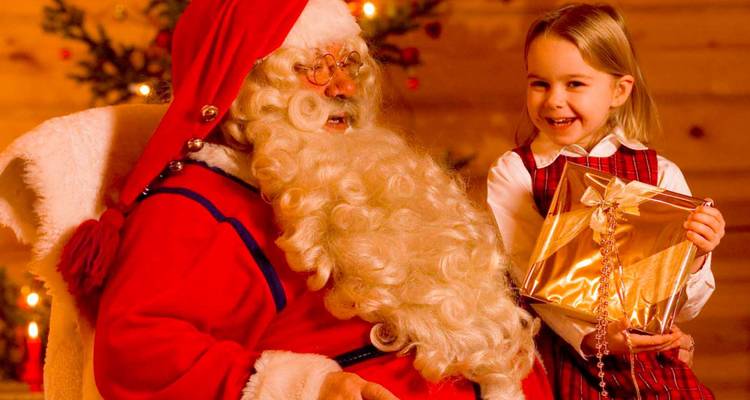 Un enfant souriant présente un cadeau emballé à une figurine du Père Noël barbu assis près d'un sapin de Noël chaleureusement illuminé.