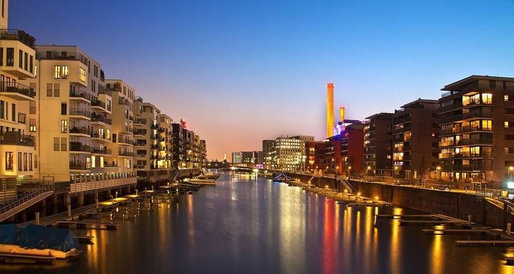 A canal flanked by modern buildings, with bright lights reflecting on the water during twilight.