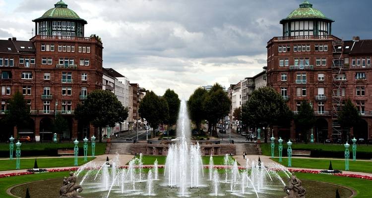 A symmetrical fountain with tall buildings in the background under a cloudy sky.