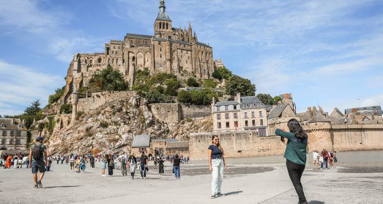 Menigtes verkennen de uitgestrekte voorplein van de Mont Saint-Michel abdij die oprijs op een rotsachtig eiland onder een blauwe hemel.