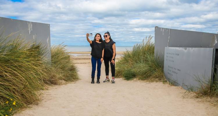 Dos mujeres tomándose una selfie en un camino arenoso que se abre hacia una playa amplia bajo un cielo parcialmente nublado.