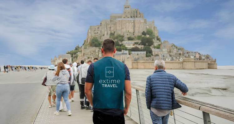 Multitud caminando por la moderna calzada hacia Mont Saint-Michel con un guía con chaleco de marca en primer plano.
