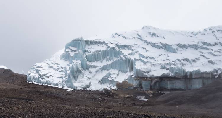 Eisklippe mit sichtbaren Gletscherspalten vor einer kargen Landschaft unter bedecktem Himmel.